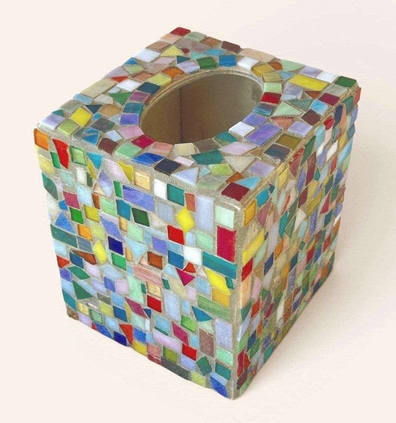 May include: A square tissue box cover featuring a multi-colored mosaic design made of small, irregular glass tiles in shades of red, blue, green, yellow, and white, set in light-colored grout with a large oval opening on the top surface.