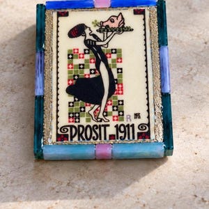 May include: A rectangular decorative tile framed in blue, purple, and pink glass pieces. The center features a graphic of a person in a black dress holding a pig head on a platter, with a checkered background and the text PROSIT 1911.
