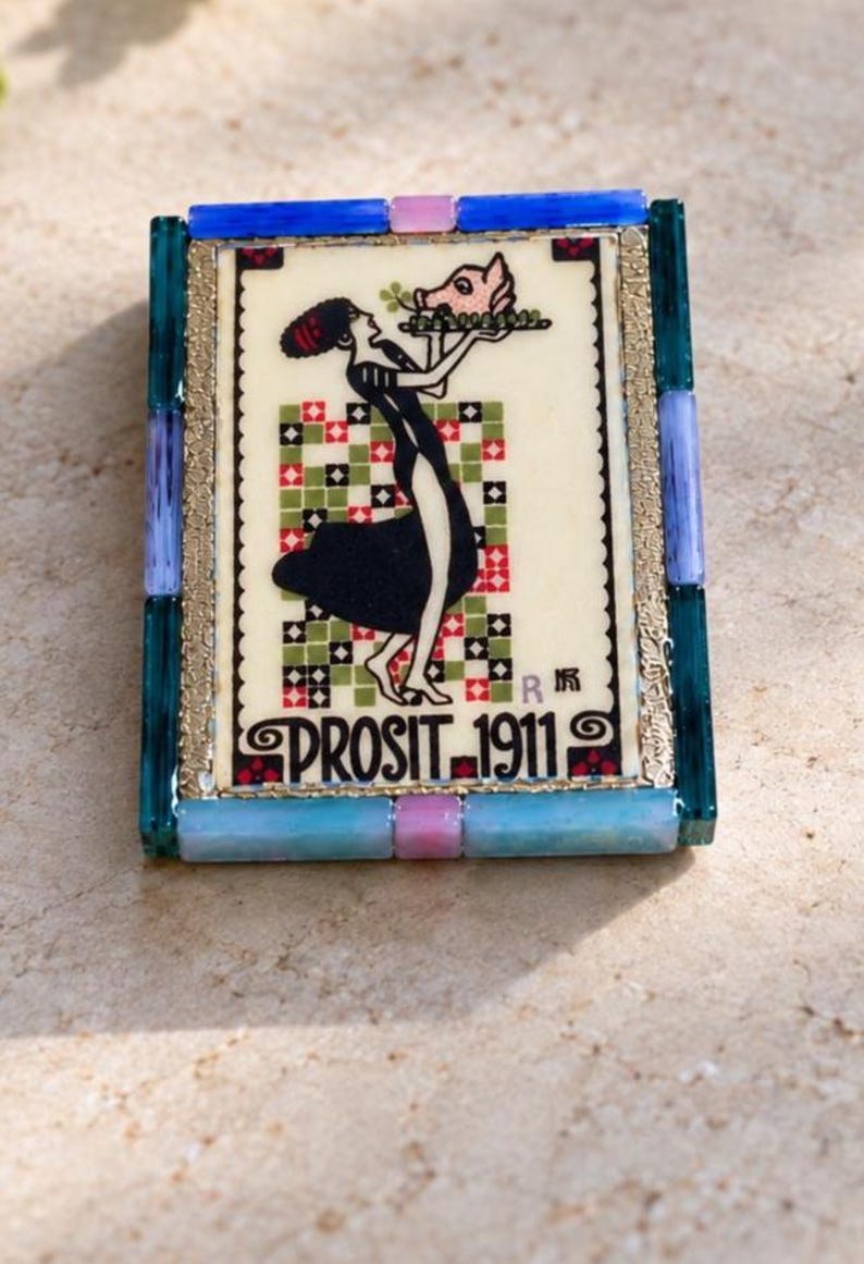 May include: A rectangular decorative tile framed in blue, purple, and pink glass pieces. The center features a graphic of a person in a black dress holding a pig head on a platter, with a checkered background and the text PROSIT 1911.