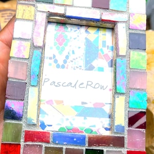 May include: A rectangular picture frame decorated with a mosaic of small, multi-colored glass tiles in shades of red, blue, green, pink, and yellow. The center of the frame contains a white insert with the handwritten text PascaleRow in black ink.