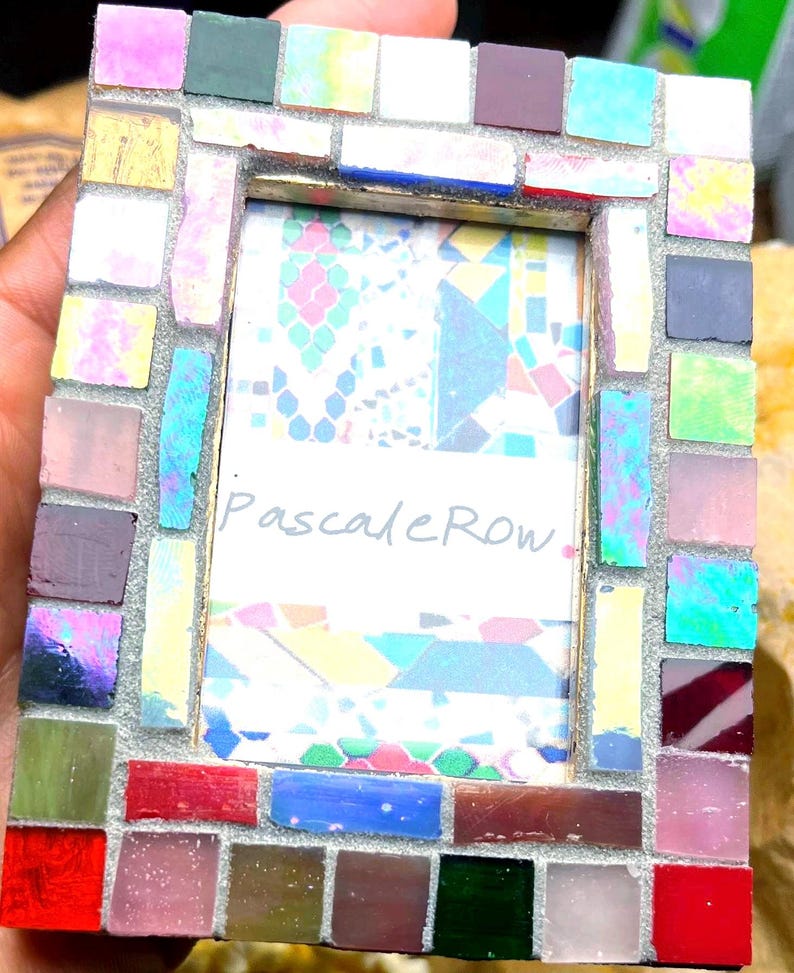 May include: A rectangular picture frame decorated with a mosaic of small, multi-colored glass tiles in shades of red, blue, green, pink, and yellow. The center of the frame contains a white insert with the handwritten text PascaleRow in black ink.