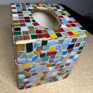 K&ouml;nnte beinhalten: Ein quadratischer Taschentuchbox-Bezug, verziert mit einem bunten Mosaik aus kleinen Glasfliesen in Rot-, Blau-, Gr&uuml;n-, Gelb- und Wei&szlig;t&ouml;nen. Die Kanten sind mit einer goldfarbenen Verzierung versehen und die Oberseite hat eine ovale &Ouml;ffnung.