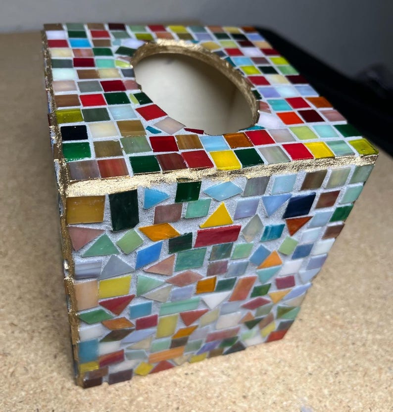 K&ouml;nnte beinhalten: Ein quadratischer Taschentuchbox-Bezug, verziert mit einem bunten Mosaik aus kleinen Glasfliesen in Rot-, Blau-, Gr&uuml;n-, Gelb- und Wei&szlig;t&ouml;nen. Die Kanten sind mit einer goldfarbenen Verzierung versehen und die Oberseite hat eine ovale &Ouml;ffnung.