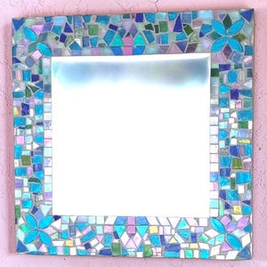 Puede incluir: Un espejo de pared cuadrado con un marco de mosaico decorativo compuesto por varias piezas de vidrio en tonos azul, verde, morado y blanco iridiscente. El marco incluye cuatro motivos florales sim&eacute;tricos hechos de vidrio turquesa brillante.