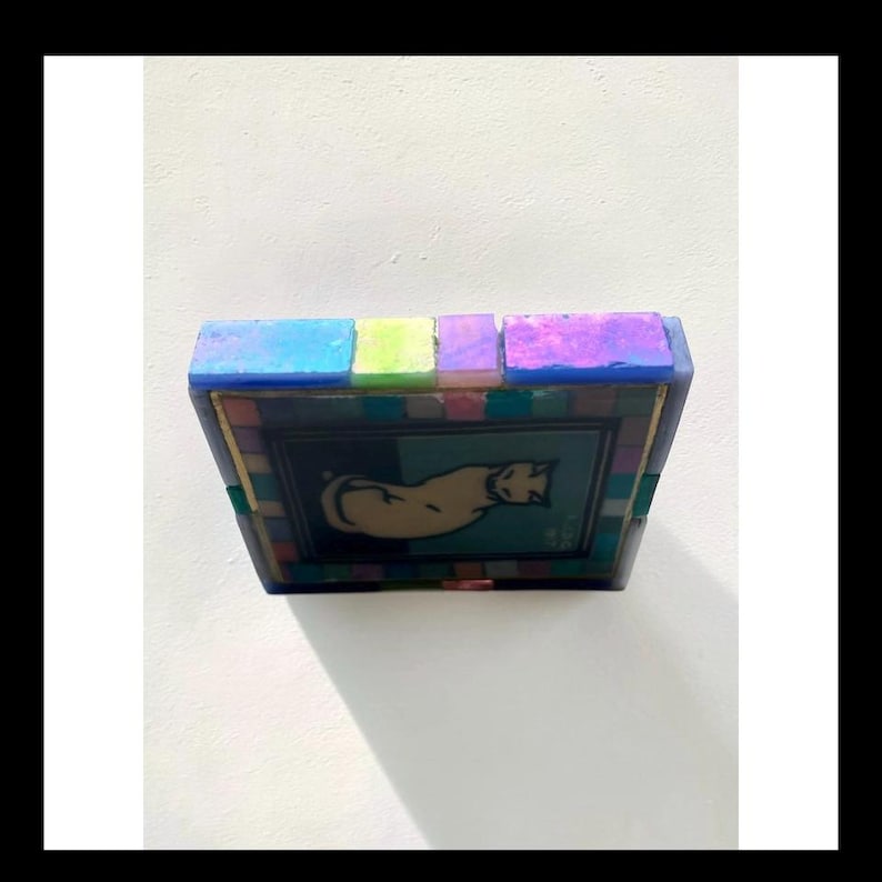 May include: A square tray featuring a central illustration of a beige cat on a dark blue background. The border is composed of small, multicolored mosaic tiles in shades of blue, pink, and green, with a top edge of larger, iridescent blue tiles.