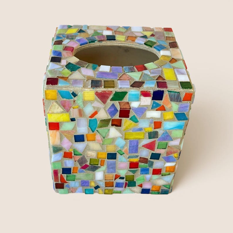 May include: A square tissue box cover featuring a multi-colored mosaic design made of small, irregularly shaped glass tiles in shades of red, blue, yellow, green, purple, and white, set into a light-colored grout with a circular opening on top.