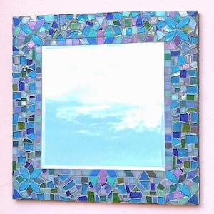 Puede incluir: Un espejo de pared cuadrado con un marco de mosaico decorativo compuesto por peque&ntilde;as piezas de vidrio de formas irregulares en tonos azul, verde, morado y turquesa, con cuatro patrones florales distintos en cada esquina del marco.