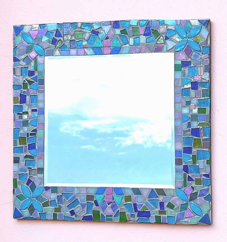 Puede incluir: Un espejo de pared cuadrado con un marco de mosaico decorativo compuesto por peque&ntilde;as piezas de vidrio de formas irregulares en tonos azul, verde, morado y turquesa, con cuatro patrones florales distintos en cada esquina del marco.