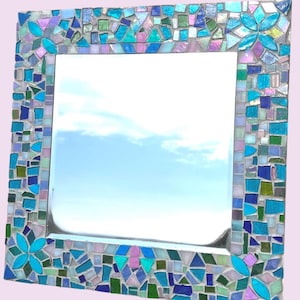 Puede incluir: Un espejo de pared cuadrado con un marco de mosaico decorativo compuesto por varias piezas de vidrio en tonos azul, turquesa, verde y violeta claro. El marco incluye cuatro detalles en forma de flor hechos con p&eacute;talos de vidrio turquesa.