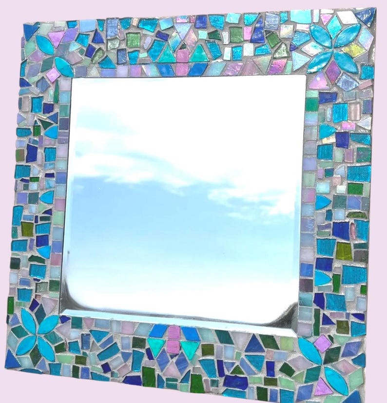 Puede incluir: Un espejo de pared cuadrado con un marco de mosaico decorativo compuesto por varias piezas de vidrio en tonos azul, turquesa, verde y violeta claro. El marco incluye cuatro detalles en forma de flor hechos con p&eacute;talos de vidrio turquesa.