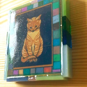 Pode incluir: Uma caixa decorativa retangular com uma ilustra&ccedil;&atilde;o central de um gato malhado laranja sobre um fundo azul escuro. A caixa &eacute; emoldurada por uma borda de pequenos azulejos de vidro multicoloridos em tons de verde, azul, roxo e rosa.