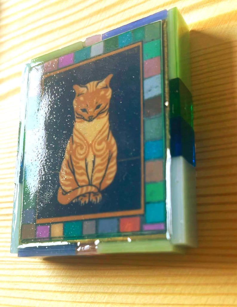 Pode incluir: Uma caixa decorativa retangular com uma ilustra&ccedil;&atilde;o central de um gato malhado laranja sobre um fundo azul escuro. A caixa &eacute; emoldurada por uma borda de pequenos azulejos de vidro multicoloridos em tons de verde, azul, roxo e rosa.