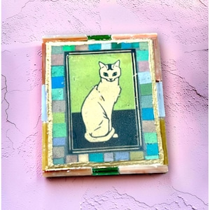 May include: A rectangular wall art piece featuring a central illustration of a seated cream-colored cat against a lime green and black background. The image is framed by a mosaic border of small, multi-colored glass tiles and gold leaf accents.