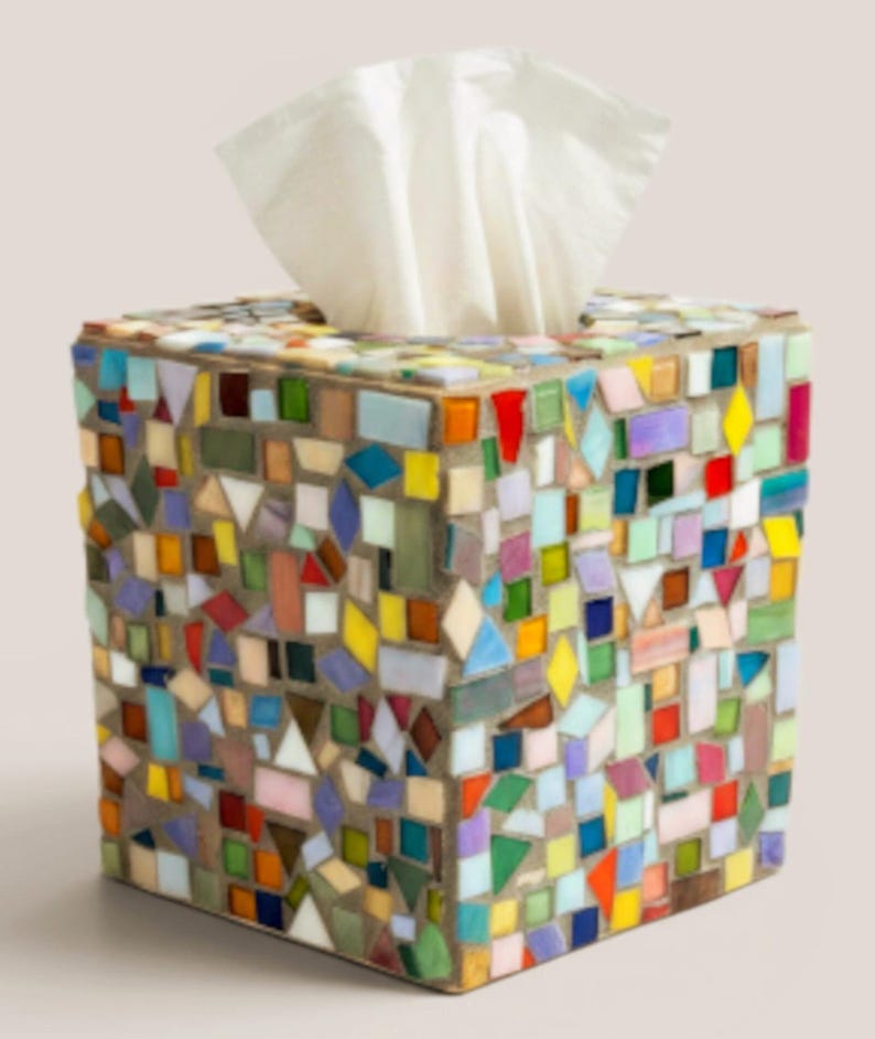 May include: A square tissue box cover decorated with a multi-colored mosaic pattern of small glass tiles in shades of red, blue, yellow, green, and purple, set in gray grout, with a white tissue protruding from the top opening of the container.