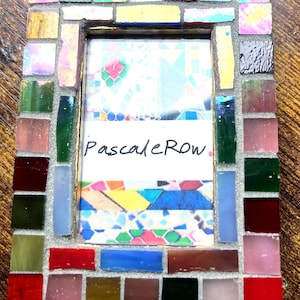 May include: A rectangular picture frame decorated with a mosaic of small, multi-colored glass tiles in shades of red, blue, green, yellow, pink, and black. The center contains a white insert with the handwritten text PascaleRow in black ink.