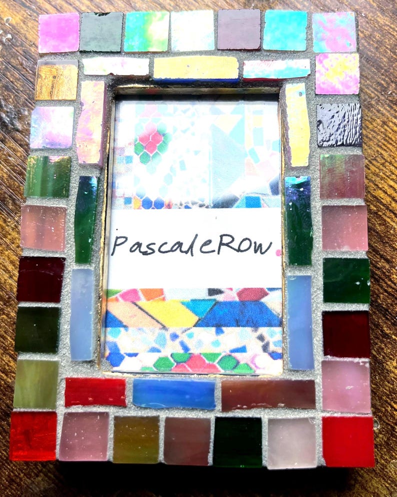 May include: A rectangular picture frame decorated with a mosaic of small, multi-colored glass tiles in shades of red, blue, green, yellow, pink, and black. The center contains a white insert with the handwritten text PascaleRow in black ink.