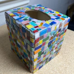 May include: A square tissue box cover decorated with a multi-colored mosaic pattern of small, irregular glass tiles in shades of blue, yellow, red, green, and pink, featuring a central oval opening on the top surface for dispensing tissues.