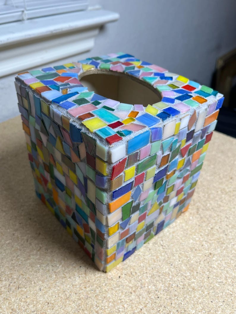 May include: A square tissue box cover decorated with a multi-colored mosaic pattern of small, irregular glass tiles in shades of blue, yellow, red, green, and pink, featuring a central oval opening on the top surface for dispensing tissues.