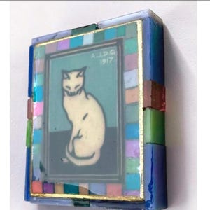 May include: A rectangular decorative object featuring a central image of a white cat sitting on a dark surface against a teal background. The text A.J.D.G 1917 is printed above the cat. The frame is composed of small, multicolored glass tiles.