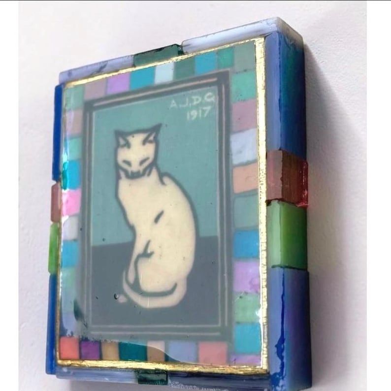 May include: A rectangular decorative object featuring a central image of a white cat sitting on a dark surface against a teal background. The text A.J.D.G 1917 is printed above the cat. The frame is composed of small, multicolored glass tiles.