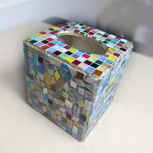 K&ouml;nnte beinhalten: Ein quadratischer Taschentuchbox-Bezug, verziert mit einem Mosaik aus kleinen, bunten Glasfliesen in Blau-, Gr&uuml;n-, Gelb-, Rot- und Wei&szlig;t&ouml;nen. Die Oberseite hat eine ovale &Ouml;ffnung und die Kanten sind mit einer strukturierten, goldfarbenen Fuge versehen.