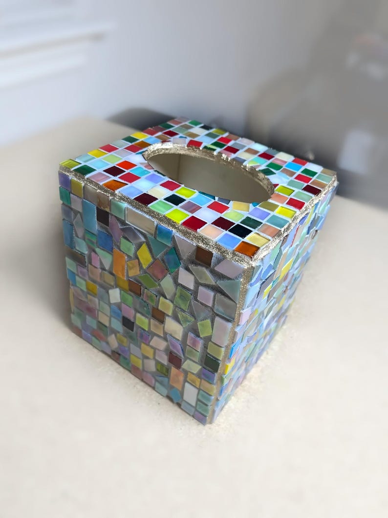 K&ouml;nnte beinhalten: Ein quadratischer Taschentuchbox-Bezug, verziert mit einem Mosaik aus kleinen, bunten Glasfliesen in Blau-, Gr&uuml;n-, Gelb-, Rot- und Wei&szlig;t&ouml;nen. Die Oberseite hat eine ovale &Ouml;ffnung und die Kanten sind mit einer strukturierten, goldfarbenen Fuge versehen.