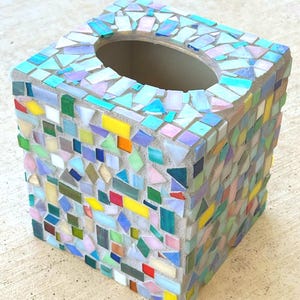 May include: A square tissue box cover decorated with a mosaic of multicolored glass tiles in various shapes and sizes. The tiles feature shades of blue, green, yellow, pink, and iridescent finishes set into a light gray grout base.