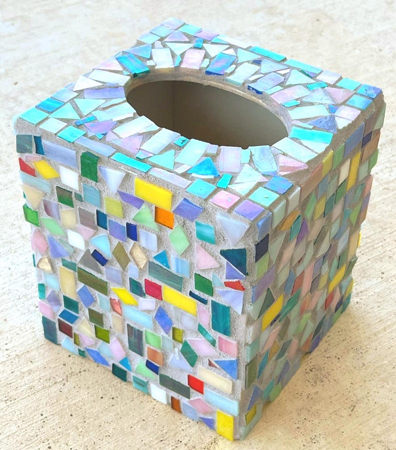 May include: A square tissue box cover decorated with a mosaic of multicolored glass tiles in various shapes and sizes. The tiles feature shades of blue, green, yellow, pink, and iridescent finishes set into a light gray grout base.