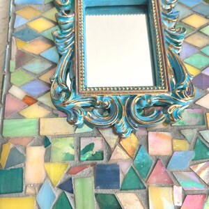 May include: A rectangular mirror featuring a central ornate frame in turquoise and gold. The surrounding border is composed of multi-colored glass mosaic tiles in shades of yellow, green, blue, pink, and white, set into a gray grout base.