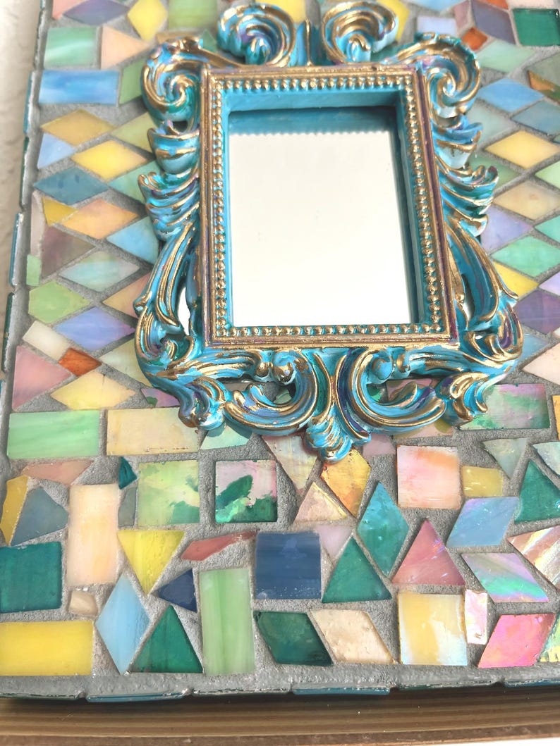 May include: A rectangular mirror featuring a central ornate frame in turquoise and gold. The surrounding border is composed of multi-colored glass mosaic tiles in shades of yellow, green, blue, pink, and white, set into a gray grout base.