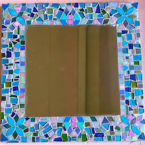 Puede incluir: Un espejo de pared cuadrado con un marco de mosaico decorativo compuesto por varios azulejos de vidrio en tonos azul, turquesa, verde y morado claro. El marco incluye cuatro patrones en forma de flor en las esquinas y los bordes.