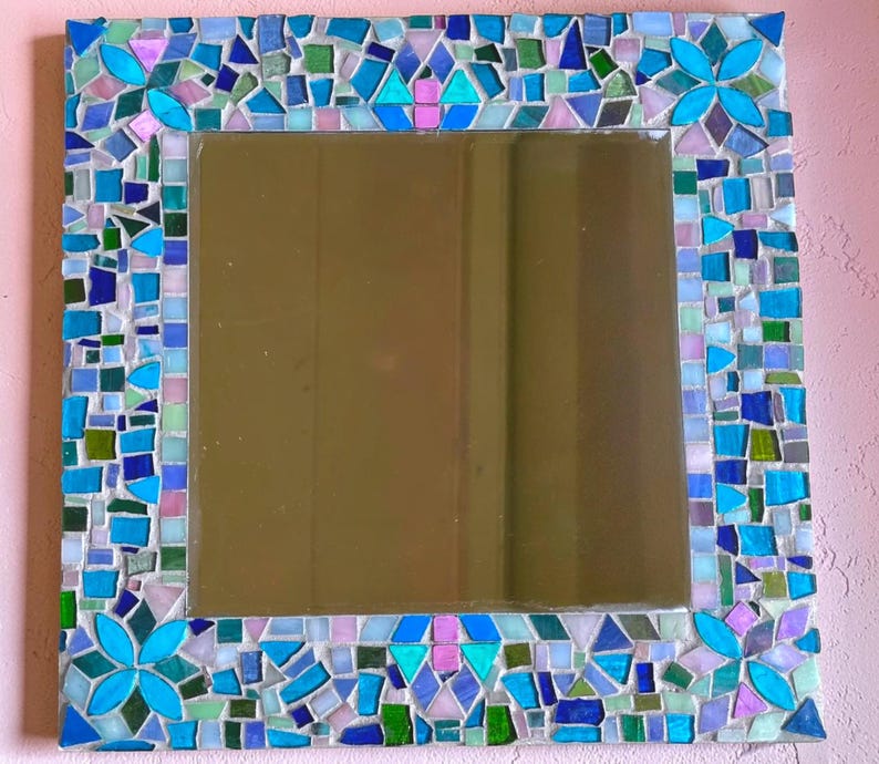 Puede incluir: Un espejo de pared cuadrado con un marco de mosaico decorativo compuesto por varios azulejos de vidrio en tonos azul, turquesa, verde y morado claro. El marco incluye cuatro patrones en forma de flor en las esquinas y los bordes.
