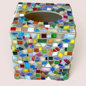 May include: A square tissue box cover decorated with a mosaic of multicolored glass tiles in shades of red, blue, green, yellow, and white. The tiles are arranged in various geometric shapes and set into a light gray grout, featuring an oval opening.