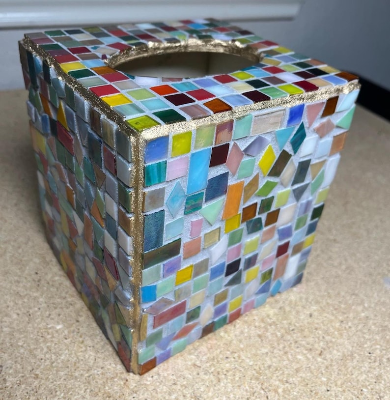 K&ouml;nnte beinhalten: Eine quadratische Taschentuchbox-Abdeckung, verziert mit einem mehrfarbigen Mosaikmuster aus kleinen Glasfliesen in Rot-, Gelb-, Blau-, Gr&uuml;n- und Rosat&ouml;nen. Die Kanten und die kreisf&ouml;rmige obere &Ouml;ffnung sind mit einer strukturierten goldfarbenen Umrandung versehen.