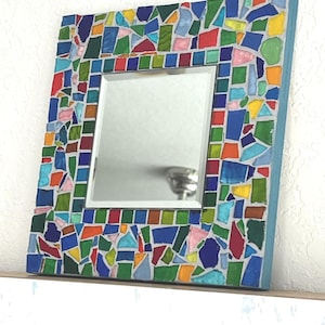 May include: A square wall mirror featuring a wide frame decorated with a colorful mosaic of irregular glass pieces in shades of blue, green, red, yellow, and pink, set against a light gray grout, with a smooth reflective center panel.