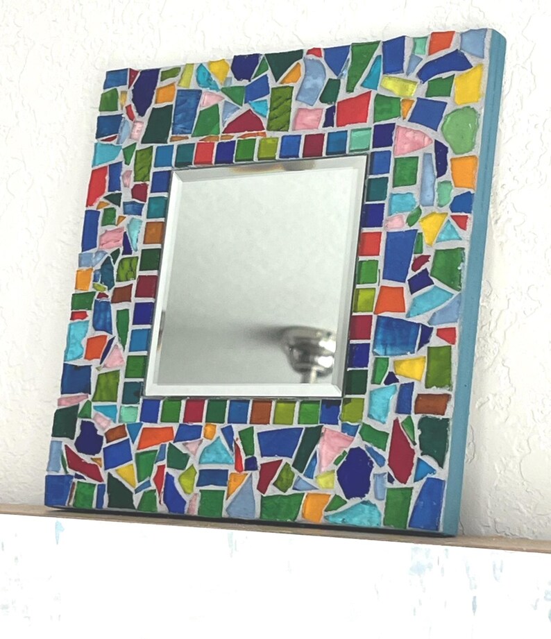 May include: A square wall mirror featuring a wide frame decorated with a colorful mosaic of irregular glass pieces in shades of blue, green, red, yellow, and pink, set against a light gray grout, with a smooth reflective center panel.