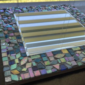 Op de afbeelding: Een vierkante spiegel met een decoratieve rand bestaande uit kleine, veelkleurige moza&iuml;ekglastegels. De tegels vertonen iriserende, blauwe, groene, roze en paarse tinten met een reflecterende afwerking, geplaatst in een lichtgekleurde voegbasis.