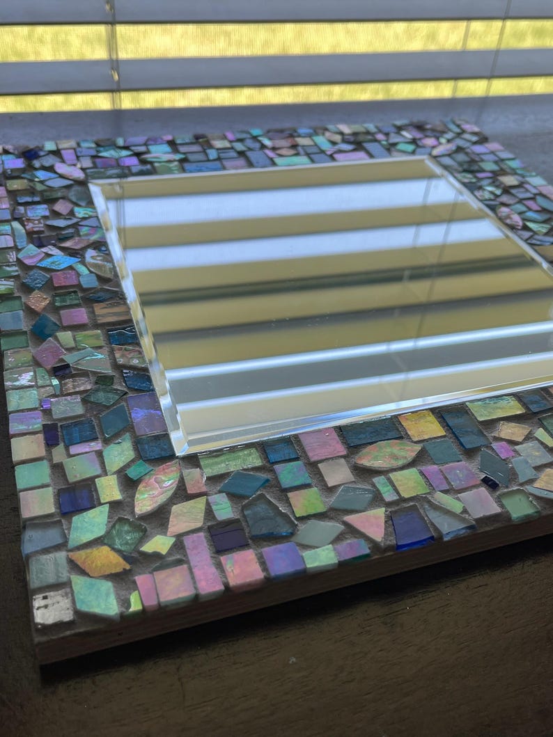 Op de afbeelding: Een vierkante spiegel met een decoratieve rand bestaande uit kleine, veelkleurige moza&iuml;ekglastegels. De tegels vertonen iriserende, blauwe, groene, roze en paarse tinten met een reflecterende afwerking, geplaatst in een lichtgekleurde voegbasis.