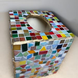 Op de afbeelding: Een vierkante tissueboxhouder versierd met een veelkleurig mozaïekpatroon van kleine glazen tegeltjes in tinten rood, geel, groen, blauw en wit. De randen zijn afgewerkt met een goudkleurige bies en de bovenkant heeft een ovale opening.