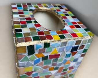 May include: A square tissue box cover decorated with a multi-colored mosaic pattern of small glass tiles in shades of red, yellow, green, blue, and white. The edges are finished with a gold-colored trim, and the top features an oval opening.