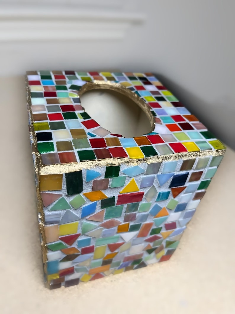 K&ouml;nnte beinhalten: Ein quadratischer Taschentuchbox-&Uuml;berzug, verziert mit einem bunten Mosaikmuster aus kleinen Glasfliesen in Rot, Gelb, Gr&uuml;n, Blau und Wei&szlig;. Die Kanten sind mit einer goldfarbenen Verzierung versehen und die Oberseite hat eine ovale &Ouml;ffnung.