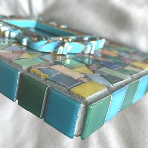 May include: A rectangular mosaic frame featuring a variety of small, multi-colored glass tiles in shades of blue, green, yellow, and pink. The center of the frame is adorned with a raised, ornate, light blue decorative border with gold accents.
