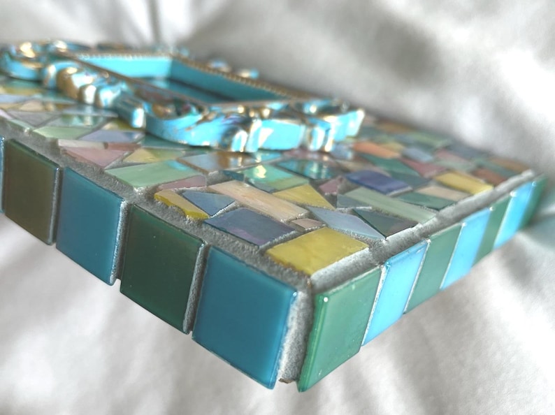 May include: A rectangular mosaic frame featuring a variety of small, multi-colored glass tiles in shades of blue, green, yellow, and pink. The center of the frame is adorned with a raised, ornate, light blue decorative border with gold accents.