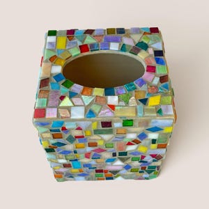 May include: A square tissue box cover featuring a multi-colored mosaic design made of small, irregular glass tiles in shades of red, blue, green, yellow, purple, and white, set into a light-colored grout with a large oval opening at the top.
