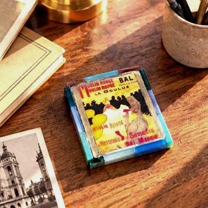 May include: A square decorative coaster featuring a vintage Moulin Rouge La Goulue poster print, framed by a border of translucent blue, green, and brown glass tiles. The central image displays yellow, black, and red text and graphics.