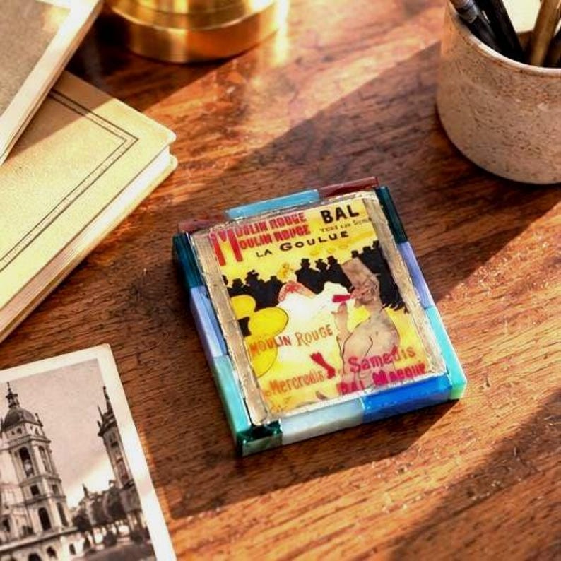 May include: A square decorative coaster featuring a vintage Moulin Rouge La Goulue poster print, framed by a border of translucent blue, green, and brown glass tiles. The central image displays yellow, black, and red text and graphics.