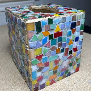 K&ouml;nnte beinhalten: Ein quadratischer Taschentuchbox-&Uuml;berzug mit einem bunten Mosaikdesign aus kleinen, unregelm&auml;&szlig;ig geformten Glasfliesen in Rot, Blau, Gr&uuml;n, Gelb und Orange. Die oberen Kanten sind mit einem goldfarbenen, metallischen Rand versehen.
