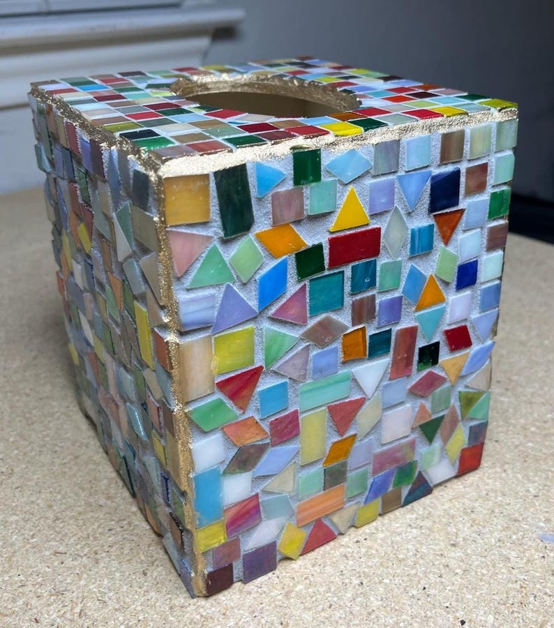 K&ouml;nnte beinhalten: Ein quadratischer Taschentuchbox-&Uuml;berzug mit einem bunten Mosaikdesign aus kleinen, unregelm&auml;&szlig;ig geformten Glasfliesen in Rot, Blau, Gr&uuml;n, Gelb und Orange. Die oberen Kanten sind mit einem goldfarbenen, metallischen Rand versehen.