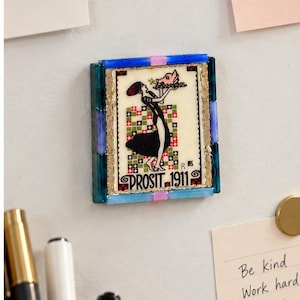 May include: A rectangular magnet featuring a vintage-style illustration of a person in a black dress holding a tray with a pink pig. The image is bordered by blue and pink glass tiles and includes the text PROSIT 1911 at the bottom.