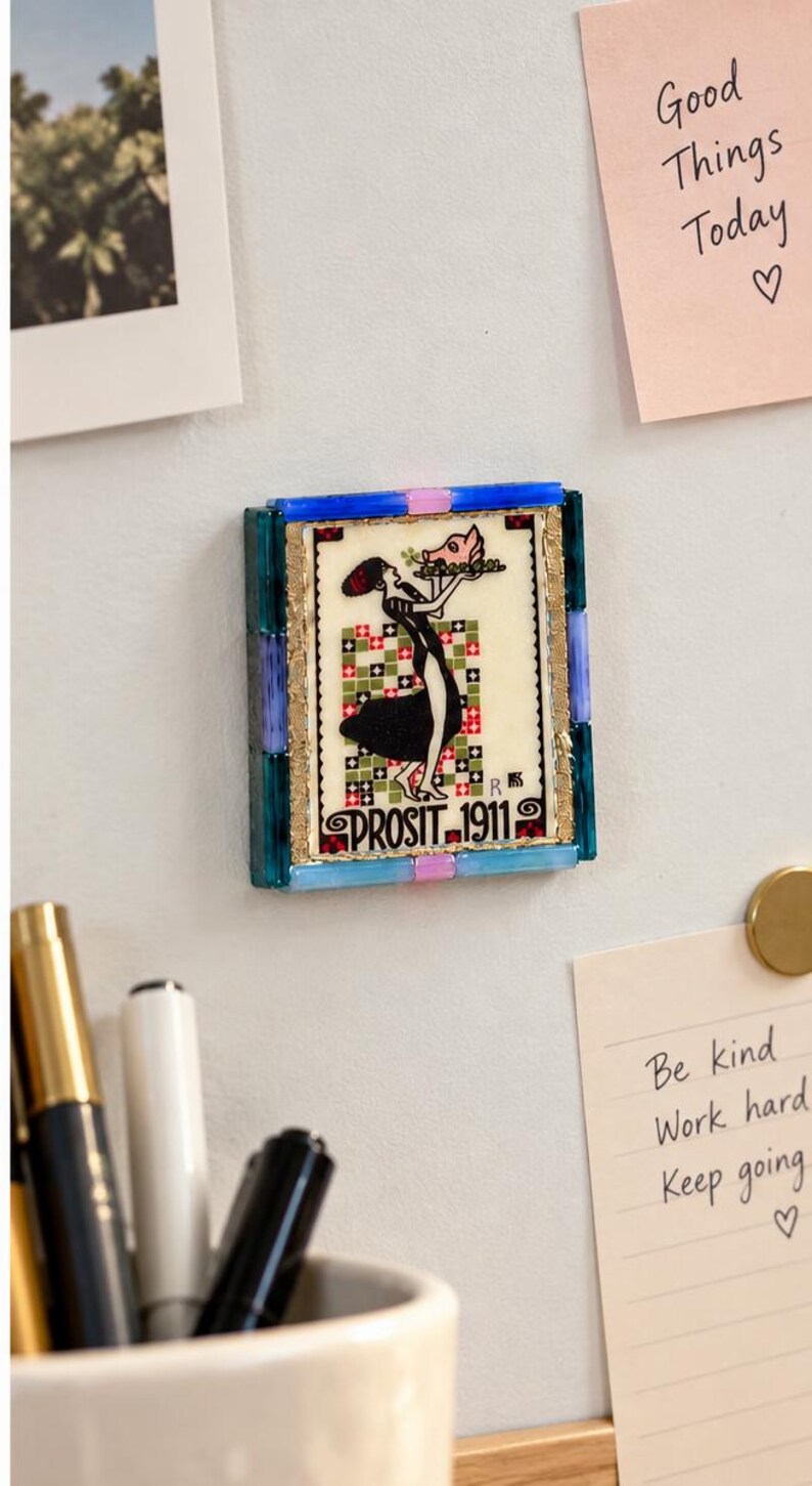 May include: A rectangular magnet featuring a vintage-style illustration of a person in a black dress holding a tray with a pink pig. The image is bordered by blue and pink glass tiles and includes the text PROSIT 1911 at the bottom.
